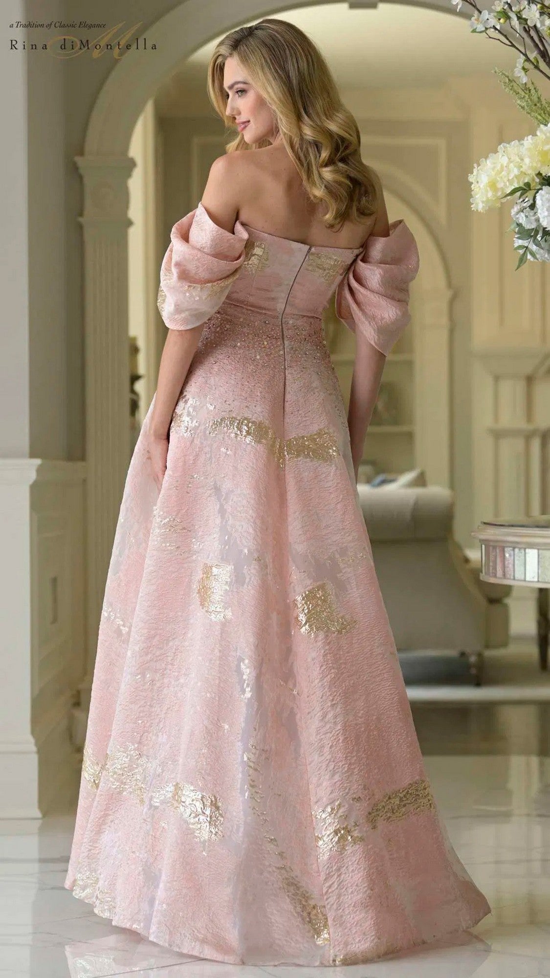 A woman stands indoors facing away, wearing the Rina Di Montella RD3405 long formal dress—an off-the-shoulder, floor-length organza gown with gold accents and an elegant a-line skirt.